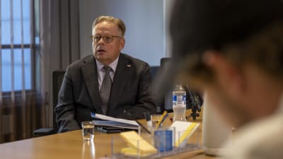 Helsingfors stads borgmästare Juhana Vartiainen sitter vid ett bord med en tunn mapp framför sig. I förgrunden skymtar journalister som deltar i den presskonferens där Vartiainen talar.