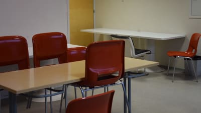 Ett beigefärgat, rektangulärt bord och röda plaststolar. I bakrunden ett vitt rektangulärt bord och flera stolar.