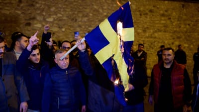 I en grupp människor står en man som håller en brinnande svensk flagga i handen. Mannens ansikte döljs nästan helt av flaggan. 