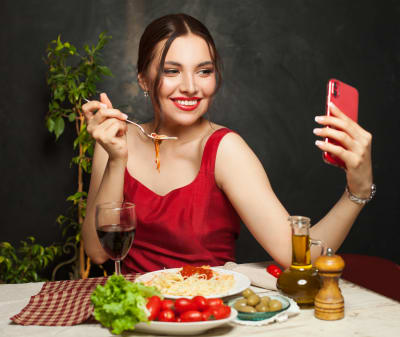 En glad kvinna äter pasta medan hon talar videosamtal med någon i telefon. 
