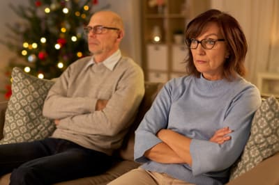 Medelålderns man och kvinna sitter i en soffa och ser sura ut, i bakgrunden en julgran