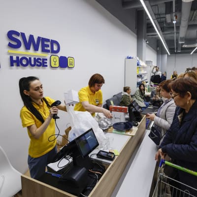 Två butiksbiträden i en kassa medan människor köar. På väggen står det "Swed house".