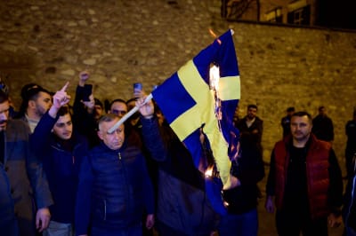 I en grupp människor står en man som håller en brinnande svensk flagga i handen. Mannens ansikte döljs nästan helt av flaggan. 