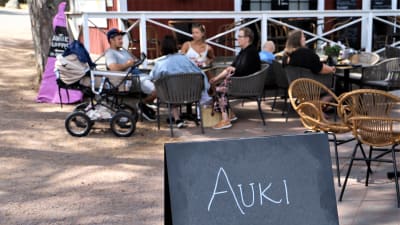 En familj med en barnvagn sitter på en uteservering i Nagu gästhamn. I förgrunden en skylt med texten "Auki".