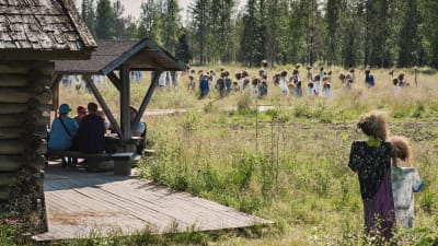 Niittykahvilassa istuvia ihmisiä (ei tunnistettavissaI, taustalla Hiljainen kansa -teoksen hahmoja.