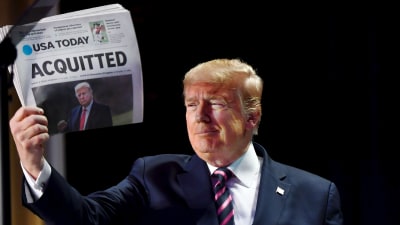 Donald Trump tittar och sidan och småler medan han håller upp en papperstidning med rubriken "frikänd" tillsammans med en bild på honom.