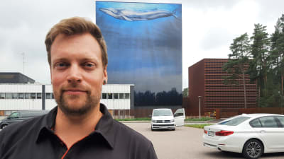 En man med kort mörkt skägg och blåsvart skjorta tittar in i kameran. I bakgrunden en blåmålad byggnad med en val på.