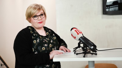 Annika Saarikko står vid ett talarpodium och talar. Framför henne mikrofoner.