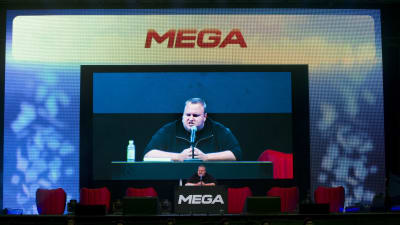 Kim Dotcom står i ett talarpodie med texten Mega. Han syns också på en stor skärm bakom podiet.