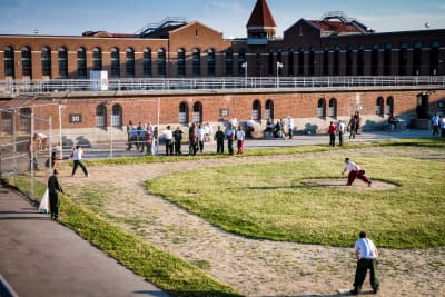 Ett tjugotal intagna står på motionsgården i Atticafängelset i USA. Några av de intagna spelar softball.