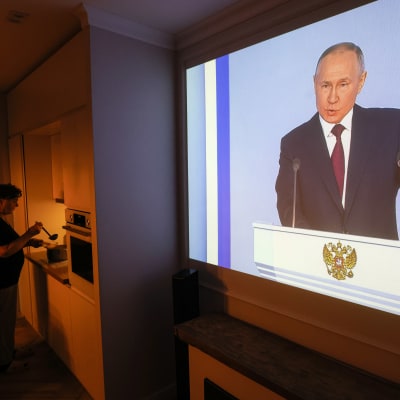 Henkilö valmistaa ruokaa keittiössä, televisioruudulla Vladimir Putin pitää puhetta.