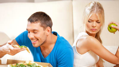 kvinna med äpple i handen tittar irriterat på glad man som äter  pizza