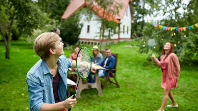En ung man och en ung kvinna spelar badminton på en gräsmatta framför ett trähus. Några personer sitter vid ett trädgårdsbord och tittar på.
