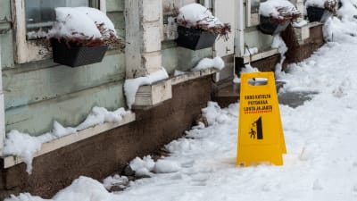En gul varningsskylt invid en husvägg varnar för fallande snö och is.