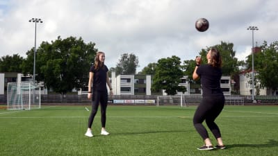 Två unga kvinnor står och sparkar en fotboll till varandra på en konstgräsplan.
