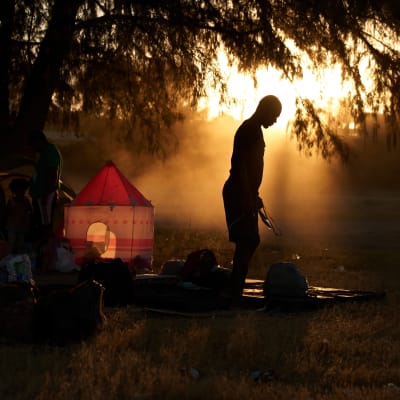 Ihminen seisoo vastavaloon ilta-aurinkoon nurmella, johon hän on laskenut isot kantamuksensa. 