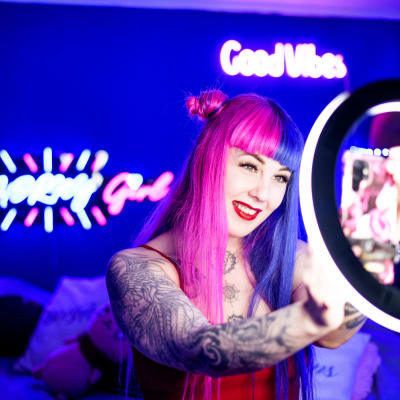 Nainen poseeraa kännykkäkameralleen huoneessa, jossa on neonvaloja ja kuvausvalo. 