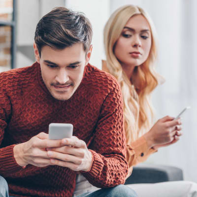 Småleende man ser på sin mobil medan en kvinna misstänksamt ser över hans axel 