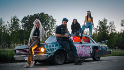 Huvudpersonerna i serien Pilsnerraggarna poserar vid en gammal bil som är svart och dekorerad med olika ord, i bakgrunden syns ett sommarlandskap.