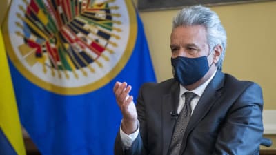 Bild på man i kostym. Han sitter med en hand upplyft och bär munskydd. I bakgrunden skymtar Ecuadors flagga.