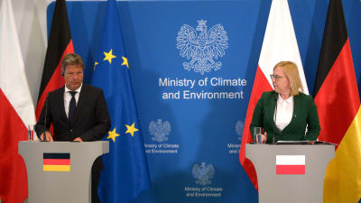 En man står bakom ett podium med en tysk flagga och en kvinna bakom ett podium med en polsk flagga. I mitten en blå vägg med texten "Ministry of Climate and Environment".