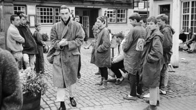 Paul Weller ulkona kävelee kohti kameraa pitkässä takissa, nuoriso katselee. Kuva dokumenttielokuvasta.