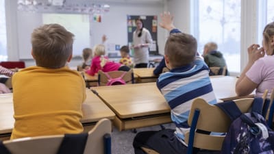 En elev sträcker upp handen och markerar under en lektion i skolan. Bredvid honom sitter en annan elev och i bakgrunden syns läraren, tavlan och fler elever.