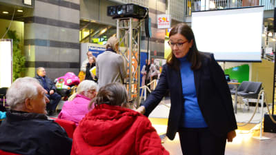 En medelålders kvinna står och intervjuar äldre personer som sitter på stolar i ett köpcentrum. Intervjuaren med ansiktet mot kameran.
