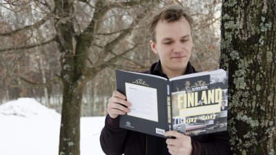 Alfred Backa som står luta mot ett träd och läser sin bok "Finland hundranånting".