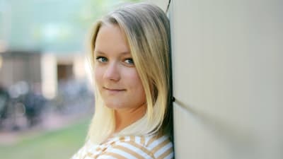 Michaela Rosenback ser in i kameran lutad mot en grå tegelvägg. Hon är klädd i en gul-vit randig t-skjort.