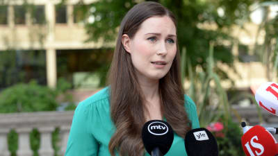 En kvinna med axellångt mörkt hår i en grön blus ser allvarlig ut och pratar mot utsträckta mikrofoner med olika mediebolags logotyper på sig.