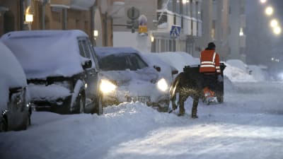 Två personer i orange västar röjer snö på en gata. En bil försöker starta. Alla bilar är täckta av snö och det snöar på bilden. Det är kväll och mörkt.