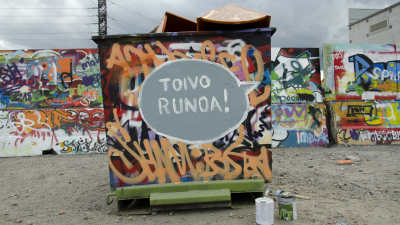 Graffiteilla maalattu roskalava, jonka päädyssä graffitina puhekupla Toivo runoa, taustalla graffiteilla maalattu betoniaita.