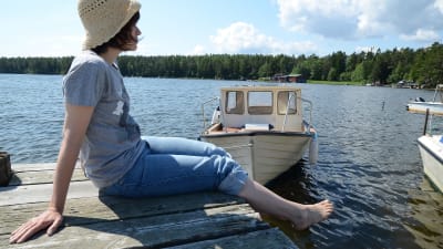 Kvinna sitter på brygga, i bakgrunden en båt.