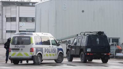 Två polisbilar utanför en grå fabrik. Ena är ett pansarfordon. 