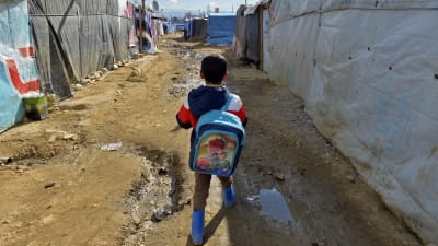 Ett syriskt flyktingbarn med blå ryggsäck promenerar i ett flyktingläger med tält i Libanon mit skolan.