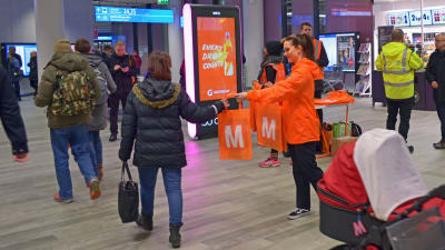 Orangeklädd personal delar ut metrokassar till passagerare i Mattby bussterminal.