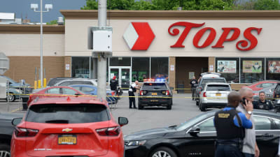 En parkeringsplats utanför en affär som heter Tops har spärrats av, en polis lutar mot en polisbil.