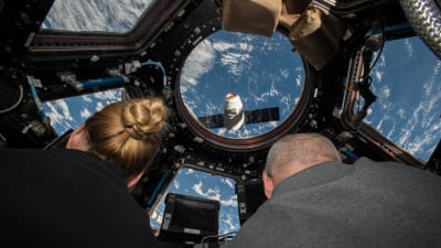 Två astronauter tittar ut från internationella rymdstationen där Dragon-leveransen närmar sig.