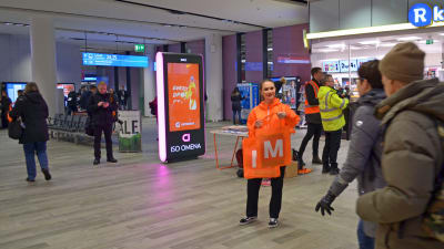 Orangeklädd personal delar ut metrokassar till passagerare i Mattby bussterminal.
