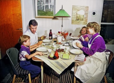 familj från 1950 talet som sitter vid ett köksbord och äter middag. Familjen ser glad och stämningen är trivsam. Modern har ett förkläde på sig. Man ser att det är hon som fixat middagen. 