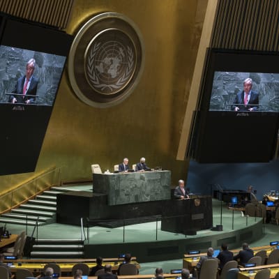 Antonio Guterres håller tal i FN:s stora sal. Han syns i talarstolen och i närbild på två stora skärmar på ömse sidor om FN-emblemet på väggen ovanför presidiet.