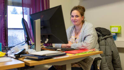 En kvinna sitter vid ett skrivbord framför en dator. Hon tittar mot kameran och ler lite.