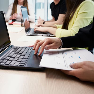 I förgrunden syns en person som studerar på datorn och har anteckningar framför sig. I bakgrunden syns fler personer som antecknar på dator eller på papper.