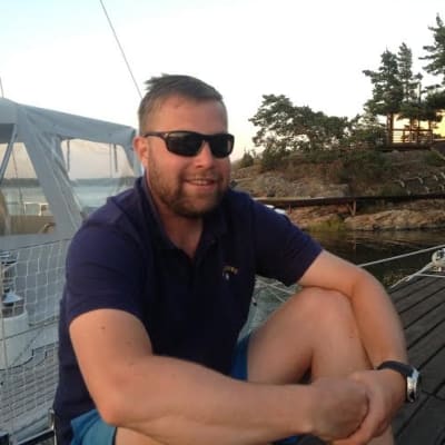 ung man sitter på brygga med bastu bakom och segelbåt bredvid sig