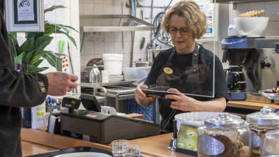 En kvinna i förkläde står bakom disken och tar betalt i en lunchrestaurang.