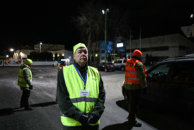En man som är strejkvakt står med gul reflexväst och gul mössa utanför en fabrik. Bakom honom står två andra personer i orange och i gul reflexväst.