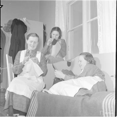Svartvit bild på tre unga kvinnor i lottadräkter som stickar i en soffa.