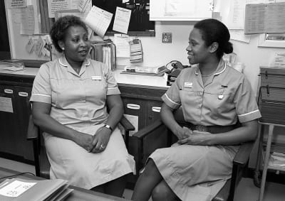Två svarta kvinnor i sjuksköterskeuniformer sitter i ett sjukhuskansli och tittar på varandra.