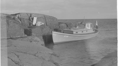 Svartvit bild: Båt är fäst vid klippa, två män lastar saker på båten.